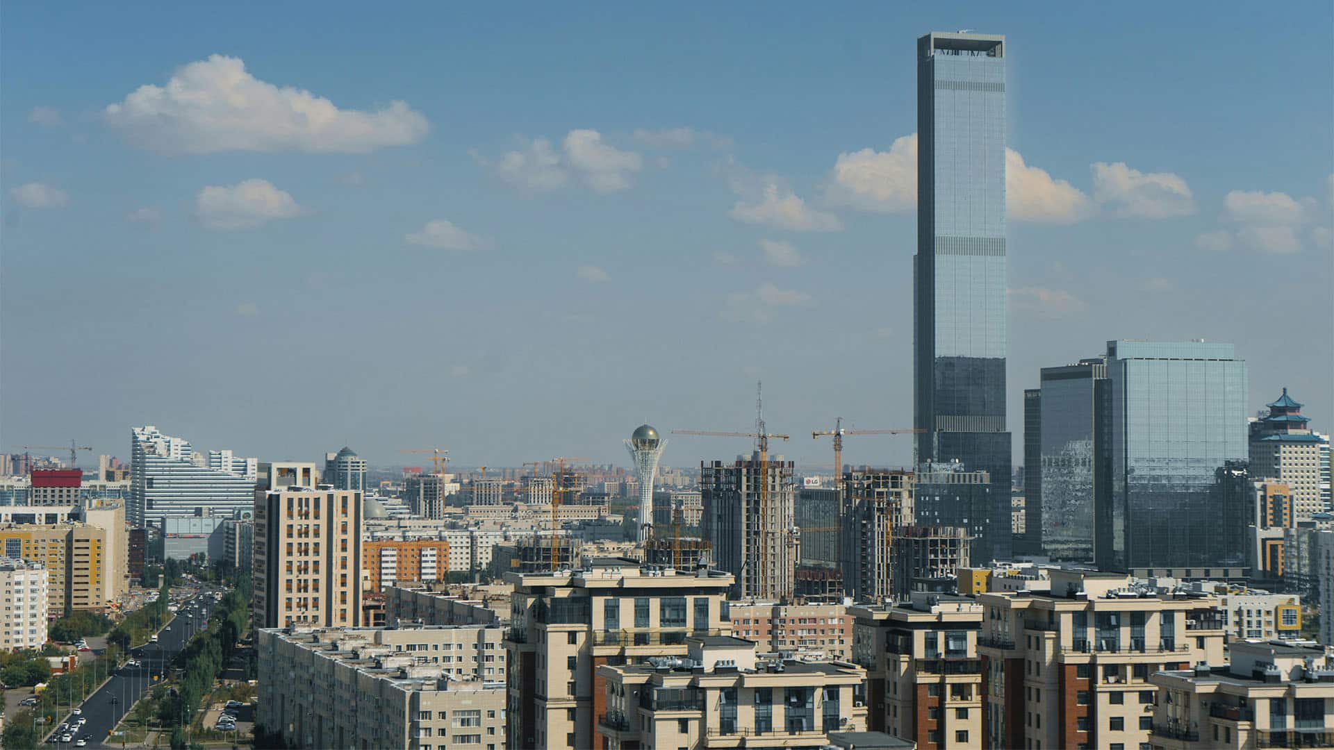 Vista aérea de la ciudad de Astaná, capital de Kazajistán. Entre el skyline, destacan edificios residenciales, y algunos de gran altura acristalados. Además, justo en el centro de la imagen, se encuentra la torre mirador de Bayterek. El cielo tiene un azul intenso, con alguna nube, aunque, en el horizonte, se vuelve más grisáceo.