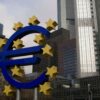 Icono del euro delante de la sede del Banco Central Europeo, en Frankfurt. El icono, de grandes dimensiones, en primer plano, deja detrás el edificio acristalado y metálico del Banco Central Europeo. Al lado del edificio, otros de color teja.