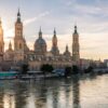 Vista de la Basílica del Pilar de Zaragoza desde el río Ebro, que recorre la ciudad. La catedral se recorta sobre el cielo azul y amarillo del atardecer. Al fondo, parte de la ciudad y uno de los puentes que la cruzan.