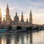 View of the Basilica del Pilar in Zaragoza from the River Ebro, which runs through the city. The cathedral stands out against the blue and yellow sky at sunset. In the background, part of the city and one of the bridges that cross it.