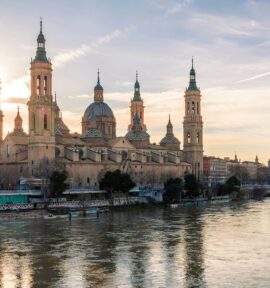 Vista de la Basílica del Pilar de Zaragoza desde el río Ebro, que recorre la ciudad. La catedral se recorta sobre el cielo azul y amarillo del atardecer. Al fondo, parte de la ciudad y uno de los puentes que la cruzan.