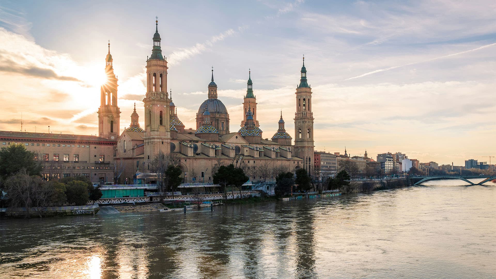 Vista de la Basílica del Pilar de Zaragoza desde el río Ebro, que recorre la ciudad. La catedral se recorta sobre el cielo azul y amarillo del atardecer. Al fondo, parte de la ciudad y uno de los puentes que la cruzan.