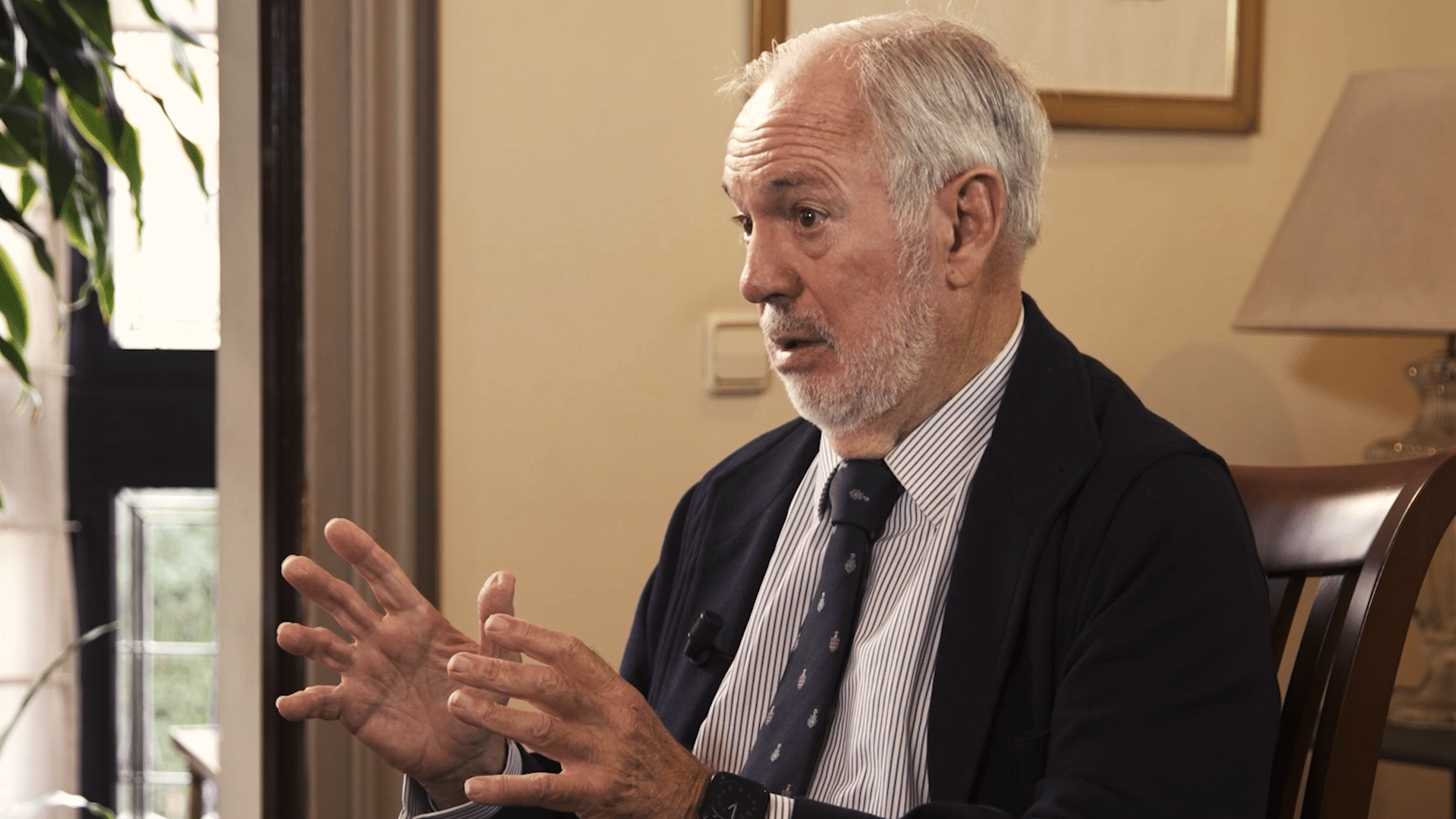 Miguel Arias Cañete conversa enfatizando con sus manos en la grabación del vídeo por los 40 años de España en la Unión Europea en la sede del Real Instituto Elcano. Arias Cañete está sentado delante de una ventana y al lado de un marco de color dorado, que ocupan parcialmente la imagen.