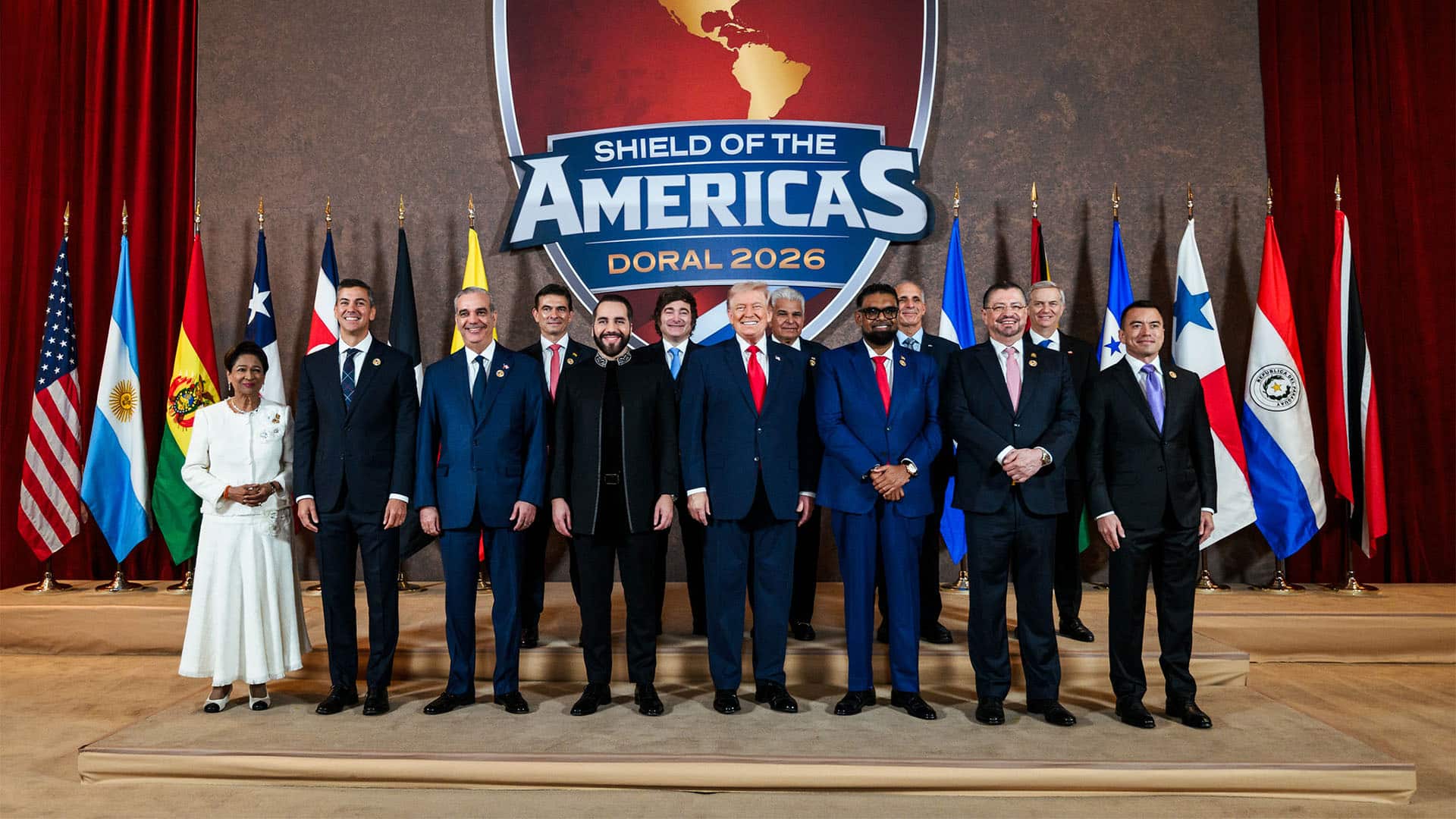 Doce representantes de los países latinoamericanos que integran Shield of the Americas posan junto al presidente de Estados Unidos en un podio de dos alturas, durante un acto oficial en el Hotel Trump National Doral, en Miami. Todos están de pie, formando dos filas ordenadas. Detrás de ellos se alinean las trece banderas de los países firmantes. En el centro del fondo destaca el logotipo de ‘Shield of the Americas’, un gran escudo de color rojo predominante que incluye el mapa del continente americano y las palabras ‘Shield of the Americas’ y ‘Doral 2026’ en letras grandes. A ambos lados del escenario cuelgan cortinas rojas.