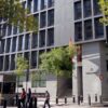 Vista general de la sede de la Audiencia Nacional, un edificio de aspecto metálico y acristalado, situado en el Paseo de la Castellana de Madrid. La fachada se ilumina con la luz del sol en un día muy despejado, y el edificio está rodeado de varios árboles. Varios transeúntes caminan por la calle, y, a la entrada del edificio, se aprecia la inscripción de la Audiencia Nacional, y el logo de la institución.