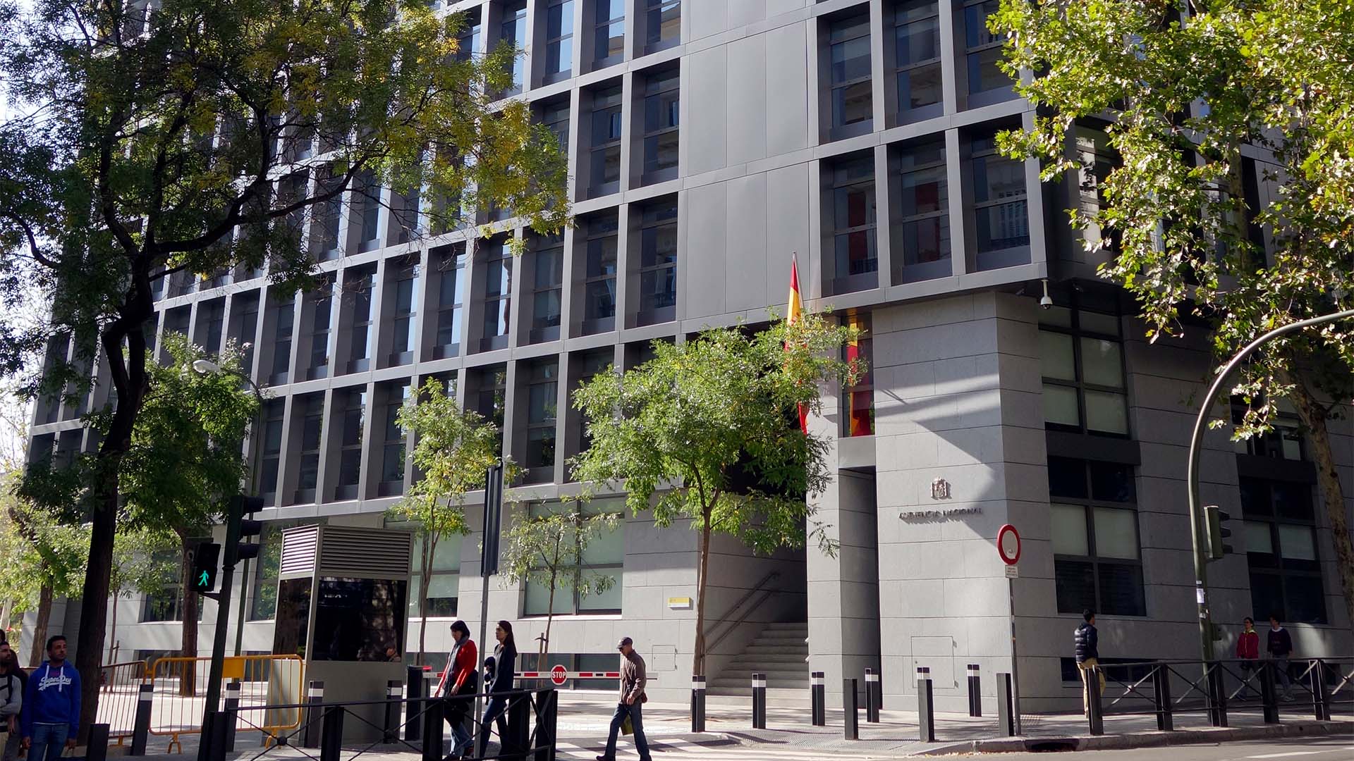 Vista general de la sede de la Audiencia Nacional, un edificio de aspecto metálico y acristalado, situado en el Paseo de la Castellana de Madrid. La fachada se ilumina con la luz del sol en un día muy despejado, y el edificio está rodeado de varios árboles. Varios transeúntes caminan por la calle, y, a la entrada del edificio, se aprecia la inscripción de la Audiencia Nacional, y el logo de la institución.