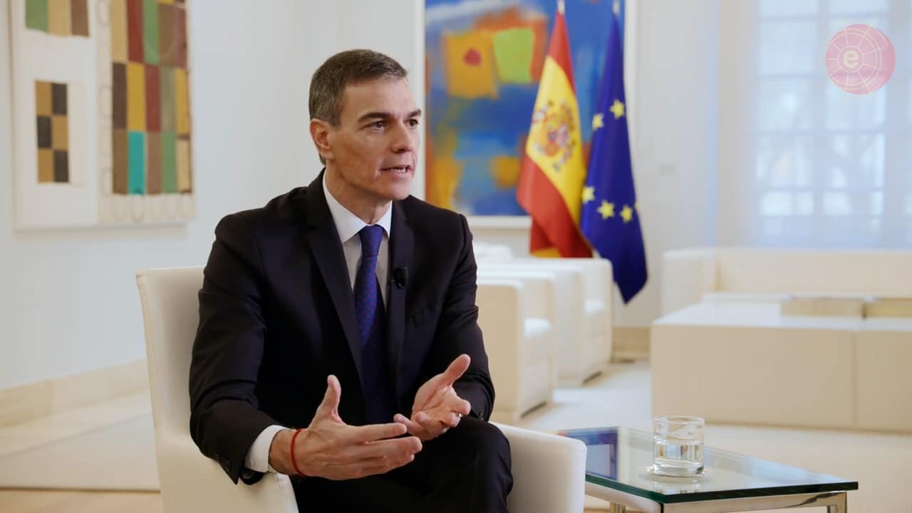 Pedro Sánchez habla en la grabación del vídeo por los 40 años de España en la Unión Europea en una de las salas del Palacio de la Moncloa. El presidente enfatiza con sus manos, sentado en una butaca de color blanco. A su lado, una mesa transparente con un vaso de agua. Al fondo, otro conjunto de butacas y mesa blancas, y, en la pared, varias obras pictóricas y, muy cerca, las banderas de España y la Unión Europea, desenfocadas.