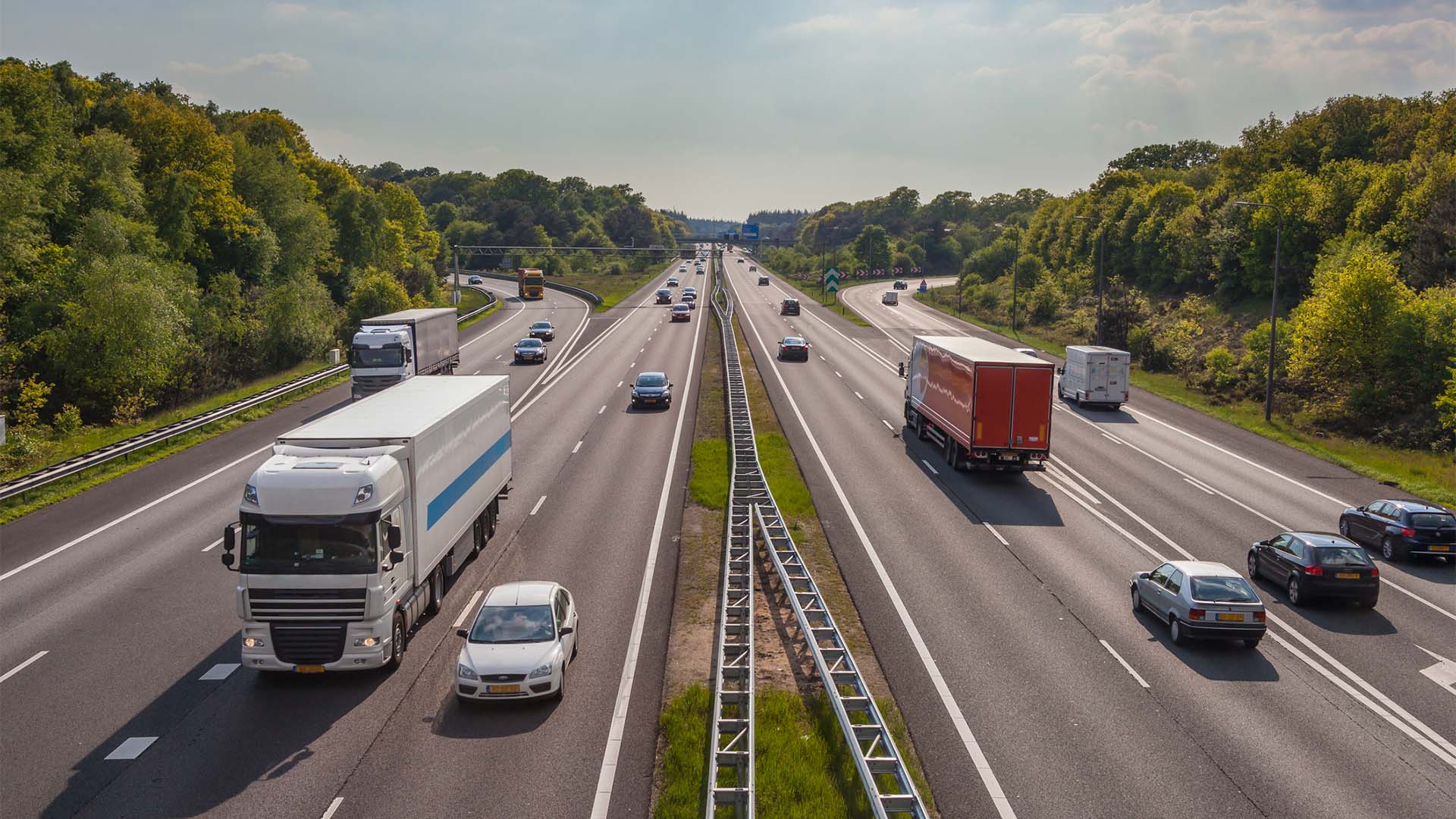 Coches y camiones circulan en ambas direcciones por la autopista A12, en Países Bajos. La mediana divide los carriles en ambos sentidos, y, a los costados, varias concentraciones de árboles. Al fondo, un cielo claro, con algunas nubes.