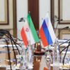 Detalle de una mesa de reunión formal con dos pequeñas banderas de Irán y Rusia colocadas en el centro rodeada de micrófonos, documentos y botellas de agua.