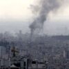 Panoramic view of Tehran as a thick column of black smoke rises from the city after an explosion, among dense buildings and construction cranes under a greyish sky.