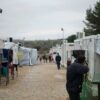 Hombre en primer plano hablando por teléfono en un albergue, con casetas blancas alineadas a ambos lados de un camino de grava y estructuras metálicas con ropa tendida a la izquierda; al fondo, varias personas caminan bajo un cielo muy nublado.