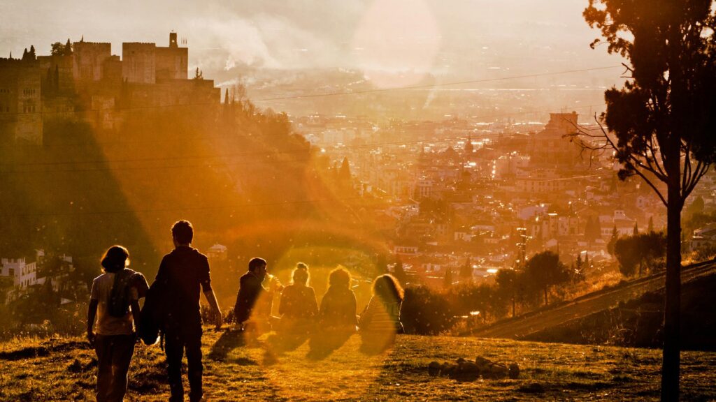 Couples and people on a hill watching the sunset in Granada (Spain). To the right is a tree, and in the background is the cityscape.